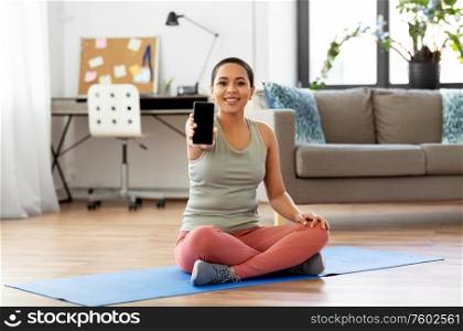 sport, fitness and technology concept - happy smiling young african american woman showing smartphone sitting on exercise mat at home. woman with smartphone sits on exercise mat at home