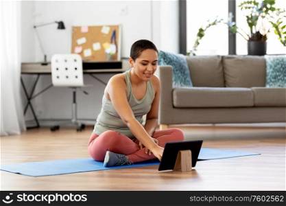 sport, fitness and technology concept - happy smiling young african american woman with tablet pc computer sitting on exercise mat at home. woman with tablet pc sits on exercise mat at home