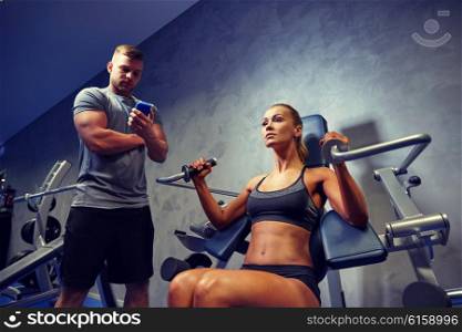 sport, fitness, bodybuilding, teamwork and people concept - young woman and personal trainer with smartphone flexing muscles on gym machine