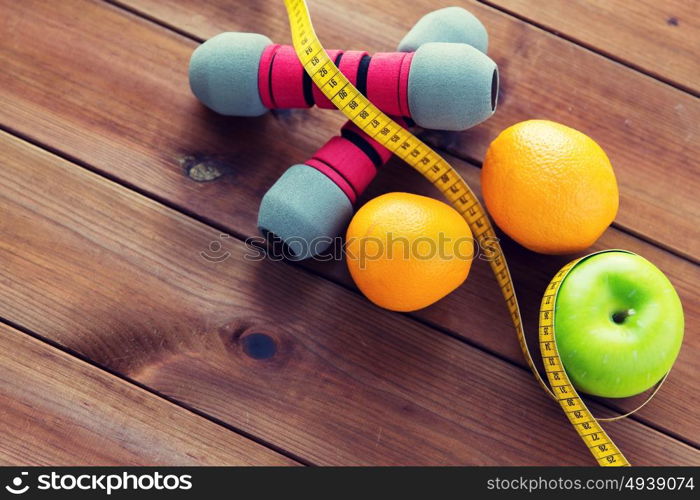 sport, fitness, diet and objects concept - close up of dumbbell and green apple with oranges wrapped by measuring tape on wooden table. close up of dumbbell, fruits and measuring tape