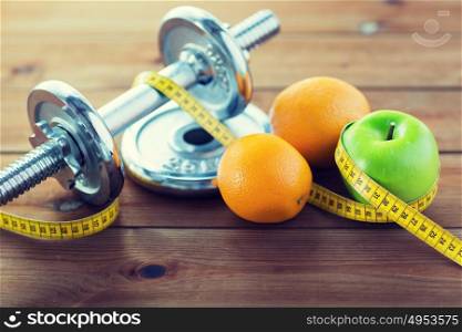 sport, fitness, diet and objects concept - close up of dumbbell and green apple with oranges wrapped by measuring tape on wooden table. close up of dumbbell, fruits and measuring tape