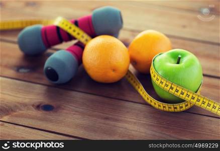 sport, fitness, diet and objects concept - close up of dumbbell and green apple with oranges wrapped by measuring tape on wooden table. close up of dumbbell, fruits and measuring tape