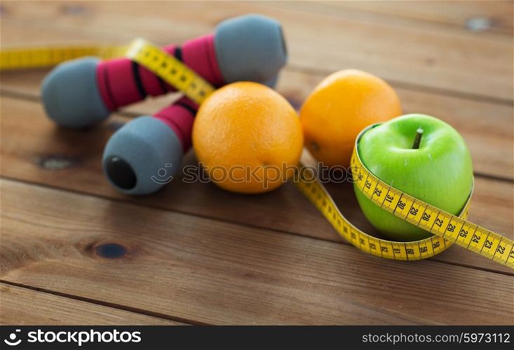 sport, fitness, diet and objects concept - close up of dumbbell and green apple with oranges wrapped by measuring tape on wooden table