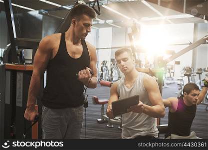 sport, fitness, equipment, technology and people concept - men with personal trainer and tablet pc computer exercising on gym machine