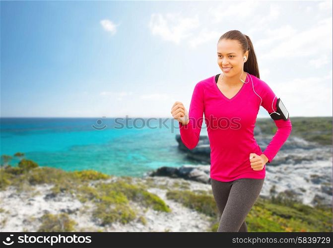 sport, fitness, health, technology and people concept - smiling young african american woman with smartphone and earphones running on beach and listening to music