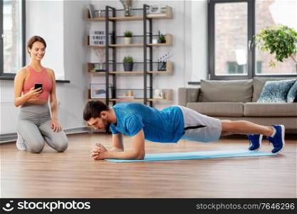 sport, fitness, lifestyle and people concept - smiling young woman with smartphone and man exercising and doing plank at home. happy couple exercising at home