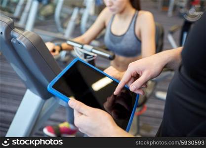 sport, fitness, lifestyle, technology and people concept - close up of trainer hands with tablet pc computer and woman working out on exercise bike in gym