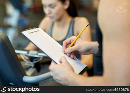 sport, fitness, lifestyle, technology and people concept - close up of trainer hands with clipboard writing and woman working out on exercise bike in gym