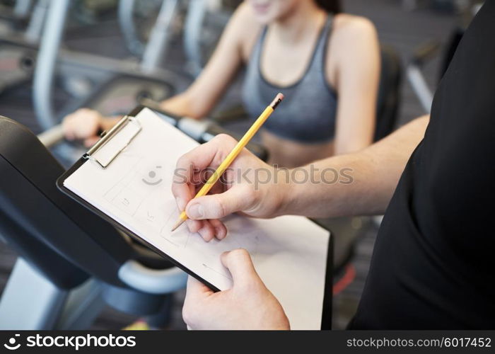 sport, fitness, lifestyle, technology and people concept - close up of trainer hands with clipboard writing and woman working out on exercise bike in gym