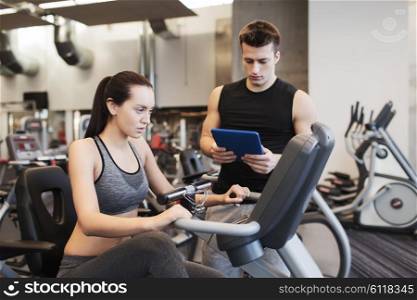sport, fitness, lifestyle, technology and people concept - woman and trainer with tablet pc computer working out on exercise bike in gym