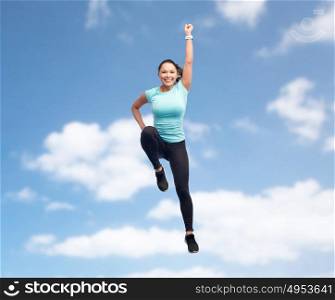 sport, fitness, motion and people concept - happy smiling young woman jumping in superhero pose over blue sky background. happy smiling sporty young woman jumping in air