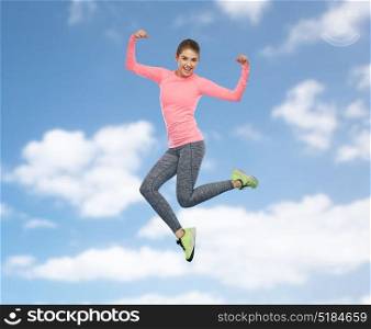 sport, fitness, motion and people concept - happy smiling young woman jumping in air and showing power gesture over blue sky and clouds background. happy smiling sporty young woman jumping in sky