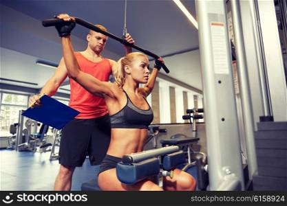 sport, fitness, teamwork and people concept - young woman flexing muscles on gym machine and personal trainer with clipboard