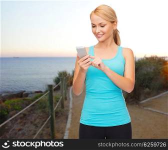 sport, fitness, technology and people concept - smiling sporty woman texting message on smartphone over beach sunset background
