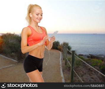 sport, fitness, technology and people concept - smiling sporty woman with smartphone and earphones listening to music over beach sunset background