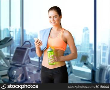 sport, fitness, technology and people concept - smiling sporty woman with smartphone, bottle of water and towel over gym machines background