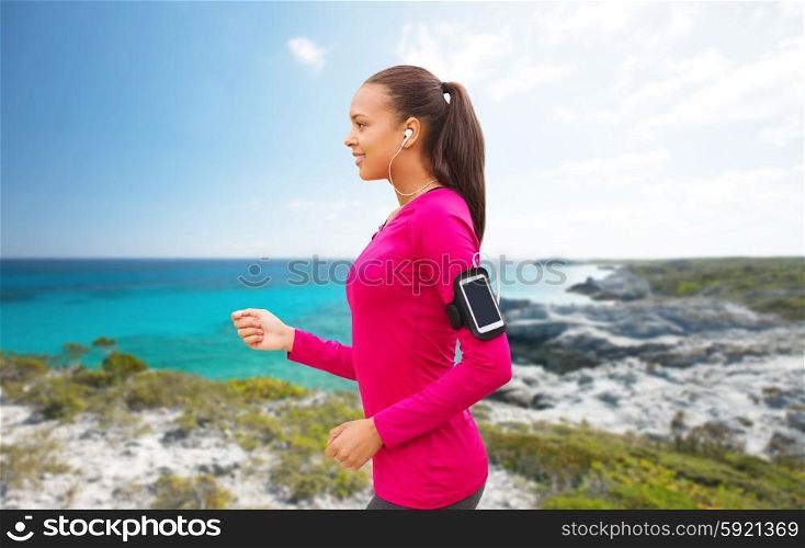 sport, fitness, technology and people concept - smiling young african american woman running with smartphone and earphones over beach background