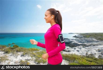 sport, fitness, technology and people concept - smiling young african american woman running with smartphone and earphones over beach background