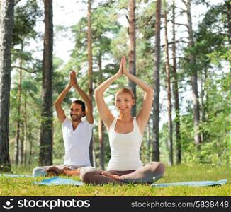 sport, fitness, yoga and people concept - smiling couple meditating and sitting on mats with raised hands over green woods background