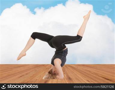 sport, fitness, yoga, people and health concept - happy young woman doing headstand exercise on wooden berth over white cloud and blue sky background