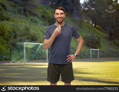 sport, football and people concept - smiling male trainer or referee with whistle over soccer field background. smiling male soccer coach or referee with whistle