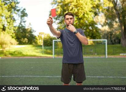 sport, football and refereeing concept - male referee whistling whistle and showing red penalty card over soccer field background. soccer referee whistling and showing red card
