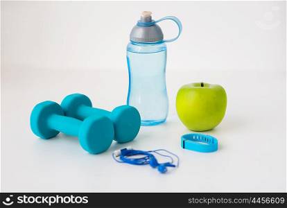 sport, healthy lifestyle and objects concept - close up of dumbbells with fitness tracker, earphones, green apple and water bottle over white background