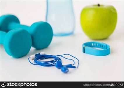 sport, healthy lifestyle and objects concept - close up of dumbbells with fitness tracker, earphones, green apple and water bottle over white background