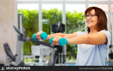 sport, healthy lifestyle and old people concept - smiling senior woman with dumbbells and fitness tracker exercising over gym background. happy old woman with dumbbells exercising in gym