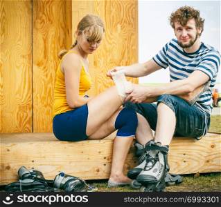 Sport injury. Couple of skaters outdoor. Young woman suffering from leg pain after taking a fall on the asphalt, man is helping bandaging her injured knee. Woman skater with injured leg knee