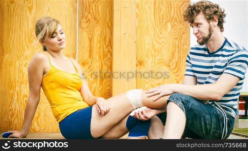 Sport injury. Couple of skaters outdoor. Young woman suffering from leg pain after taking a fall on the asphalt, man is helping bandaging her injured knee. Woman skater with injured leg knee
