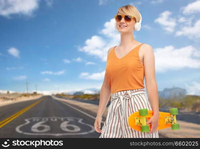 sport, leisure and skateboarding concept - smiling teenage girl in sunglasses and headphones with short modern cruiser skateboard over route 66 background. smiling teenage girl with skateboard over