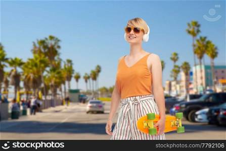 sport, leisure and skateboarding concept - smiling teenage girl in sunglasses and headphones with short modern cruiser skateboard over venice beach background in california. teenage girl with skateboard over venice beach