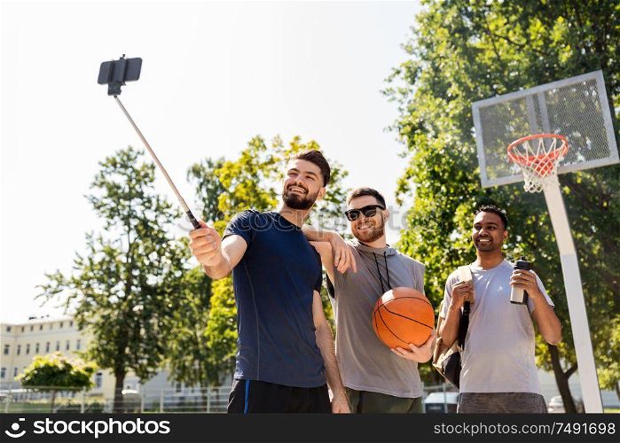 sport, leisure games and male friendship concept - group of happy men or friends taking picture by smartphone on selfie stick at outdoor basketball playground. happy men taking selfie at basketball playground