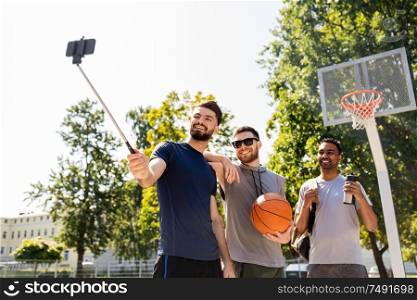 sport, leisure games and male friendship concept - group of happy men or friends taking picture by smartphone on selfie stick at outdoor basketball playground. happy men taking selfie at basketball playground