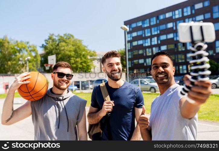 sport, leisure games and male friendship concept - group of happy men or friends taking selfie by smartphone on tripod at outdoor basketball playground. happy men taking selfie at basketball playground