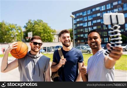 sport, leisure games and male friendship concept - group of happy men or friends taking selfie by smartphone on tripod at outdoor basketball playground. happy men taking selfie at basketball playground