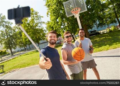 sport, leisure games and male friendship concept - group of happy men or friends taking picture by smartphone on selfie stick on outdoor basketball playground. happy men taking selfie on basketball playground