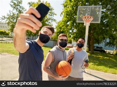 sport, leisure games and male friendship concept - group of happy men or friends wearing face protective masks for protection from virus disease taking selfie on outdoor basketball playground. happy men taking selfie on basketball playground
