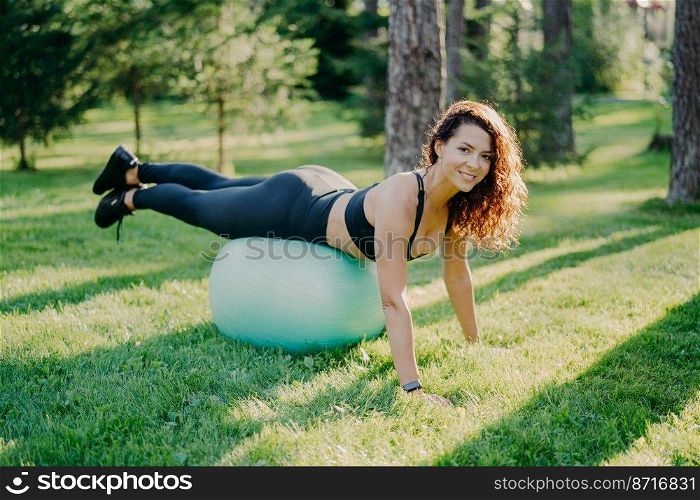 Sport, recreation, fitness and healthy lifestyle concept. Brunette sporty young European woman in active wear leans on fitness ball, makes physical exercises outdoor, enjoys favorite activity
