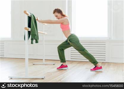 Sport, recreation, flexibility concept. Motivated strong female makes plank, leans at ballet barre, stretches before exercises prepares muscles or biceps for hard training. Aerobics instructor indoor