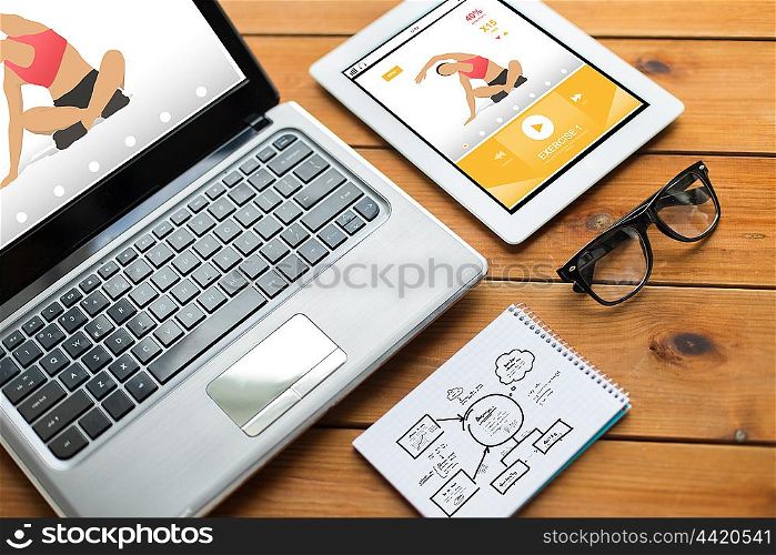 sport, responsive design and technology concept - close up of on laptop computer, tablet pc, notebook and eyeglasses with fitness application and scheme on wooden table