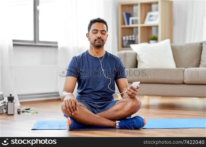 sport, technology and healthy lifestyle concept - indian man in earphones listening to music on smartphone at home. indian man with smartphone on exercise mat at home