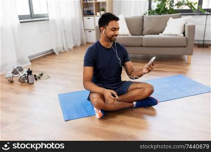 sport, technology and healthy lifestyle concept - smiling indian man in earphones listening to music on smartphone at home. indian man with smartphone on exercise mat at home