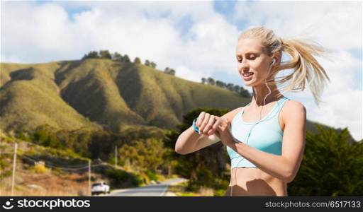sport, technology and healthy lifestyle concept - smiling young woman with fitness tracker and earphones exercising over big sur hills and road background in california. woman with fitness tracker doing sports outdoors. woman with fitness tracker doing sports outdoors