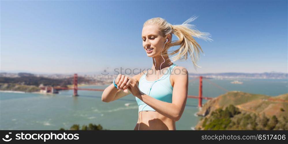 sport, technology and healthy lifestyle concept - smiling young woman with fitness tracker and earphones exercising over golden gate bridge in san francisco bay background. woman with fitness tracker over golden gate bridge. woman with fitness tracker over golden gate bridge