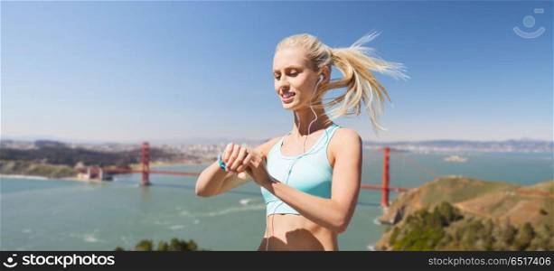 sport, technology and healthy lifestyle concept - smiling young woman with fitness tracker and earphones exercising over golden gate bridge in san francisco bay background. woman with fitness tracker over golden gate bridge. woman with fitness tracker over golden gate bridge