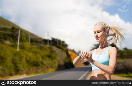 sport, technology and healthy lifestyle concept - smiling young woman with fitness tracker and earphones exercising over big sur hills and road background in california. woman with fitness tracker doing sports outdoors. woman with fitness tracker doing sports outdoors