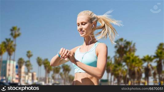 sport, technology and healthy lifestyle concept - smiling young woman with fitness tracker and earphones exercising over venice beach background in california. woman with fitness tracker doing sports. woman with fitness tracker doing sports
