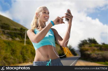 sport, technology and healthy lifestyle concept - smiling young woman with smartphone, earphones and fitness tracker listening to music over big sur hills and road background in california. woman with smartphone and earphones doing sports. woman with smartphone and earphones doing sports